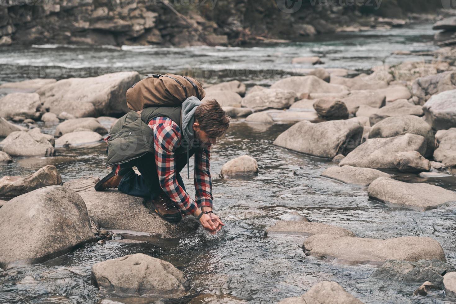 necesita un refrigerio. apuesto joven moderno bebiendo agua del río mientras camina por las montañas foto