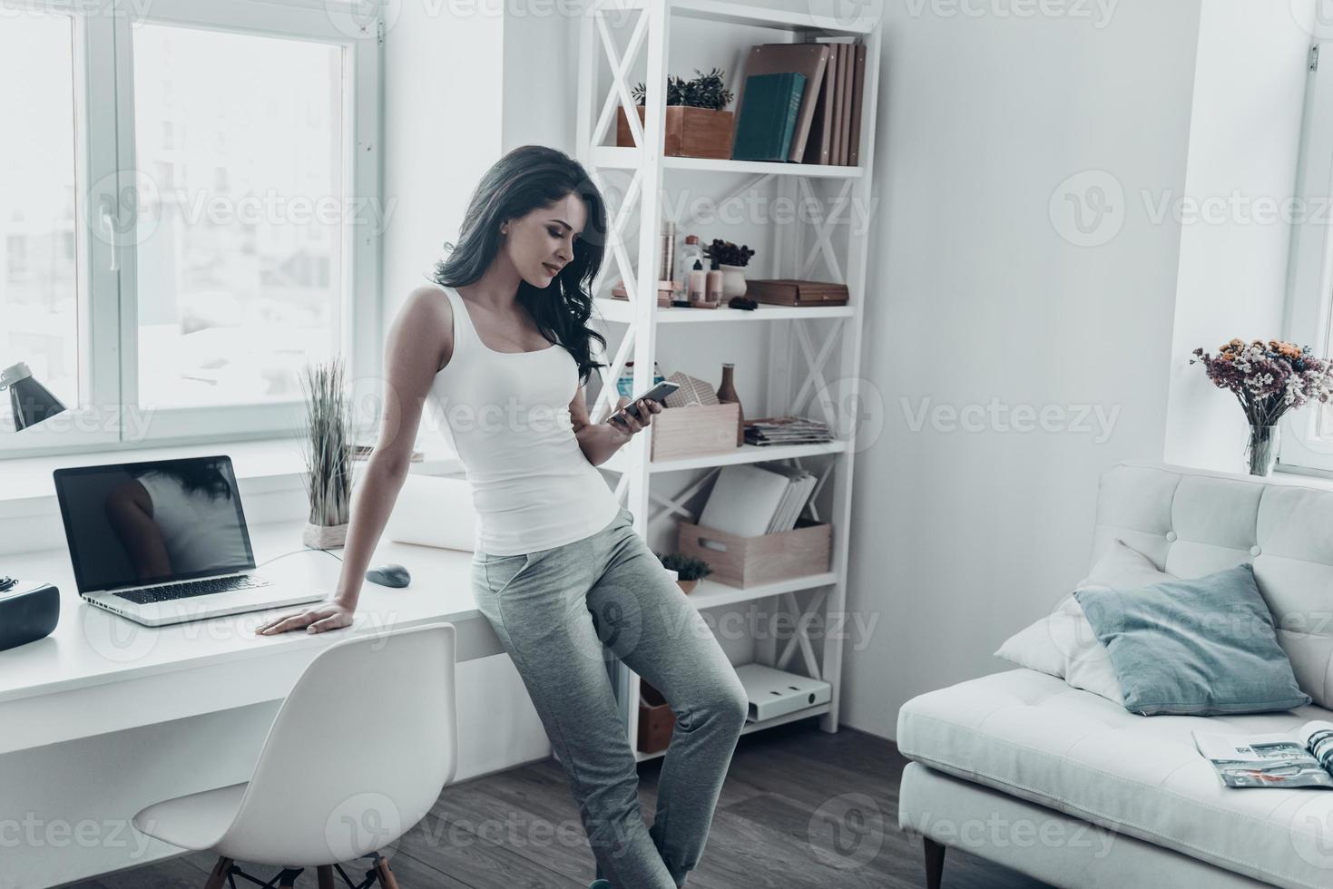 Enjoying nice chat with friend. Attractive young woman looking at mobile phone and smiling while leaning on the desk at home photo