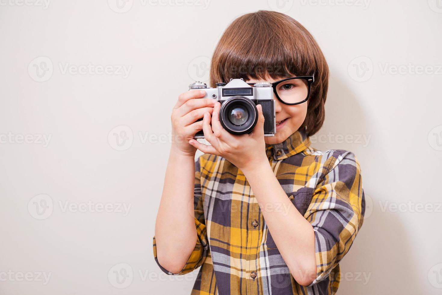 le gusta disparar. niño pequeño con anteojos sosteniendo la cámara y sonriendo mientras está de pie contra el fondo gris foto