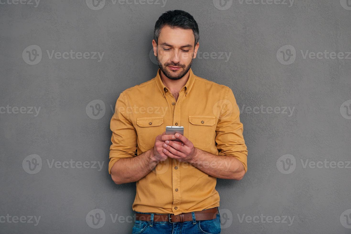 Quick message to friend. Confident mature man holding smart phone and looking at it while standing against grey background photo