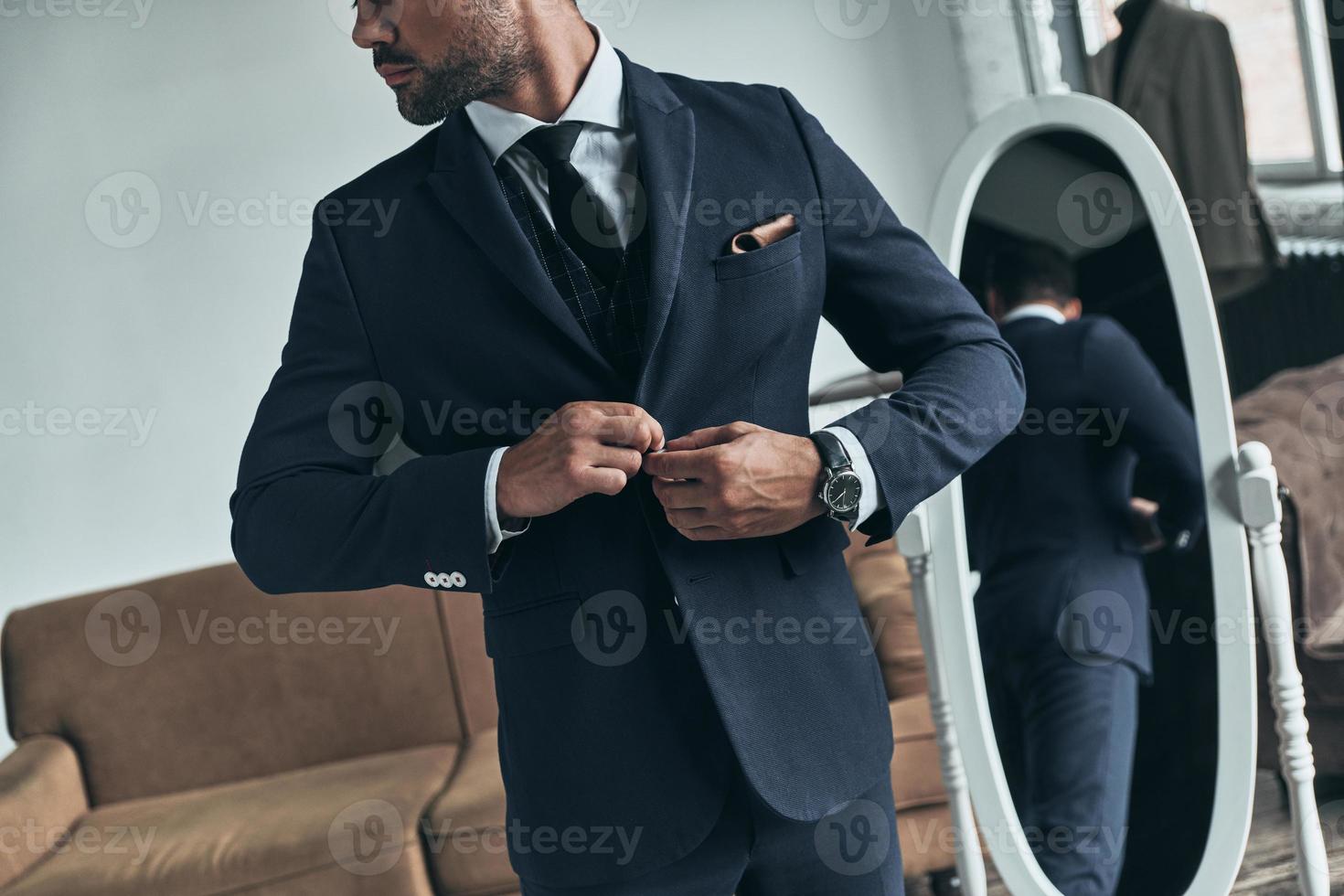 Confident in his perfect style. Close up of young man in full suit buttoning his jacket while standing indoors photo