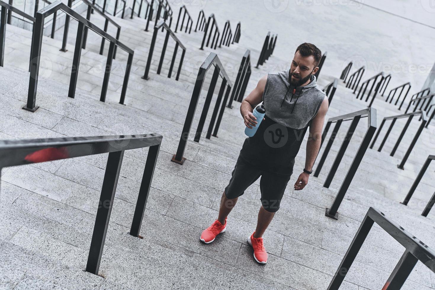 hidratarse siempre es importante. un joven apuesto con ropa deportiva sosteniendo una botella de agua mientras estaba parado en los escalones afuera foto