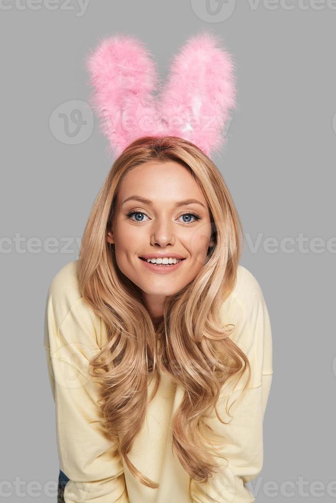 Beautiful female. Attractive young smiling woman in pink bunny ears gesturing and looking at camera while standing against grey background photo