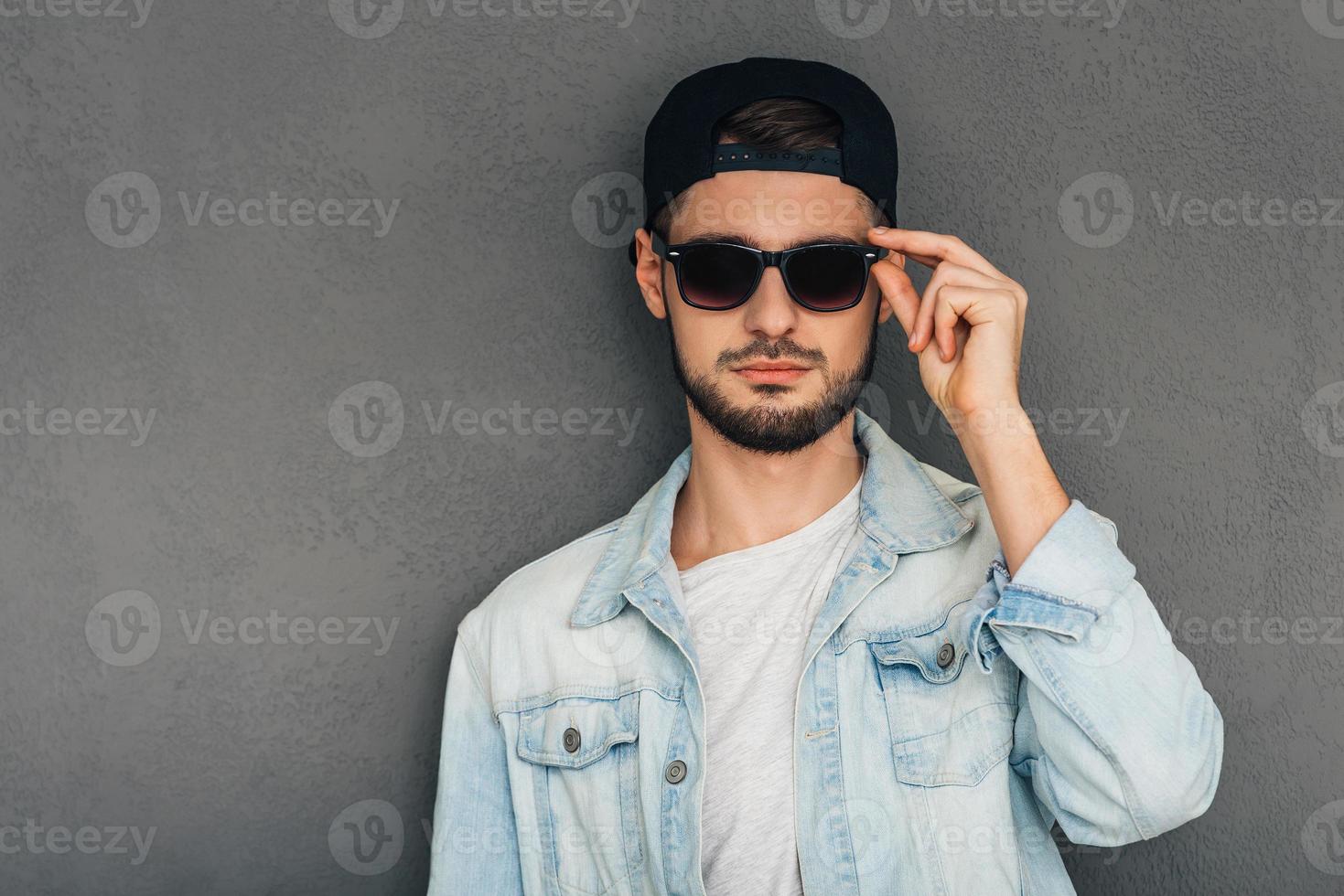 Do you like my new sunglasses Confident young man adjusting his sunglasses and looking at camera while standing against grey background photo