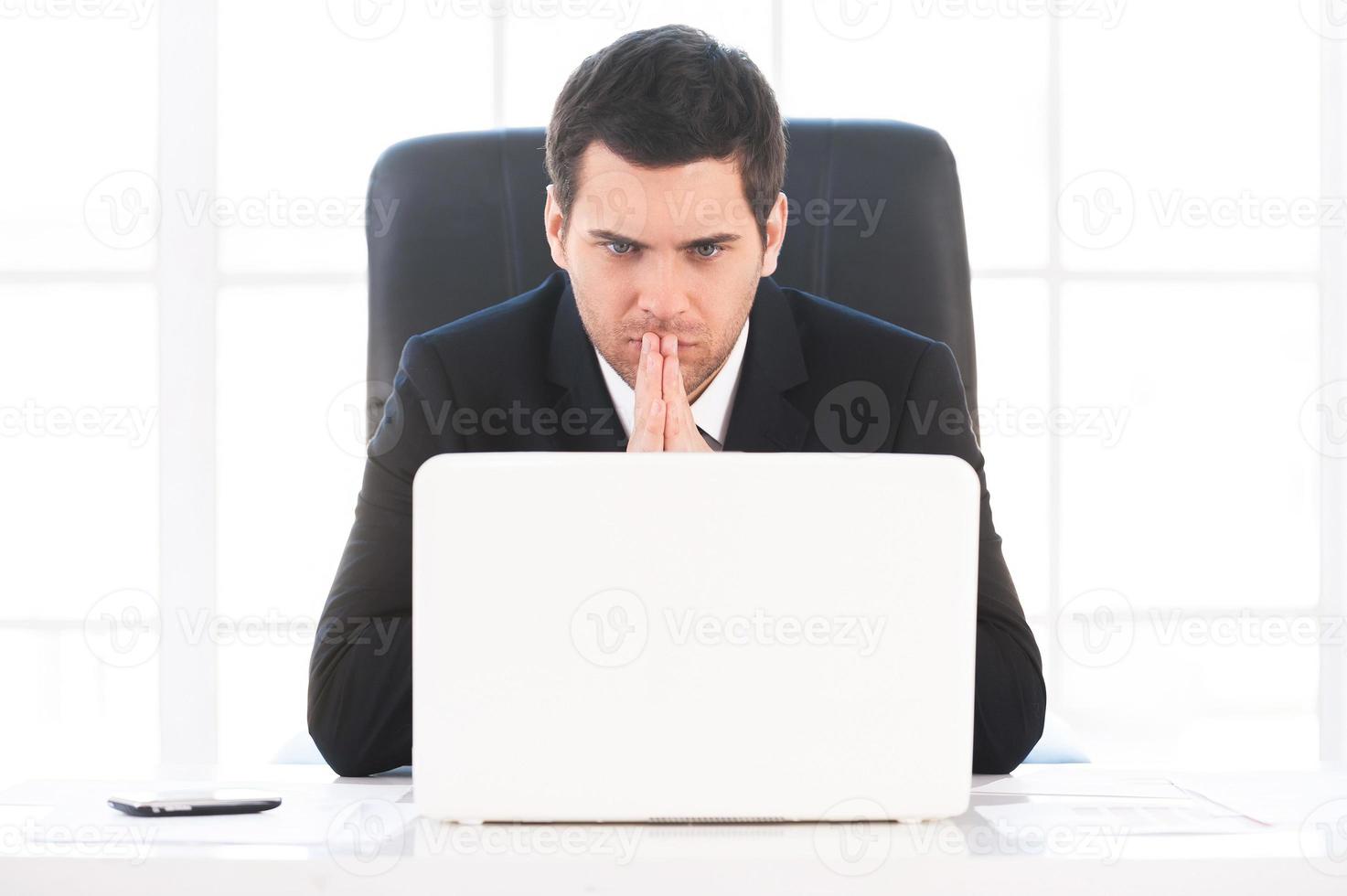 Finding out solution. Worried young man in formalwear looking at the computer and holding hands clasped while sitting at his working place photo