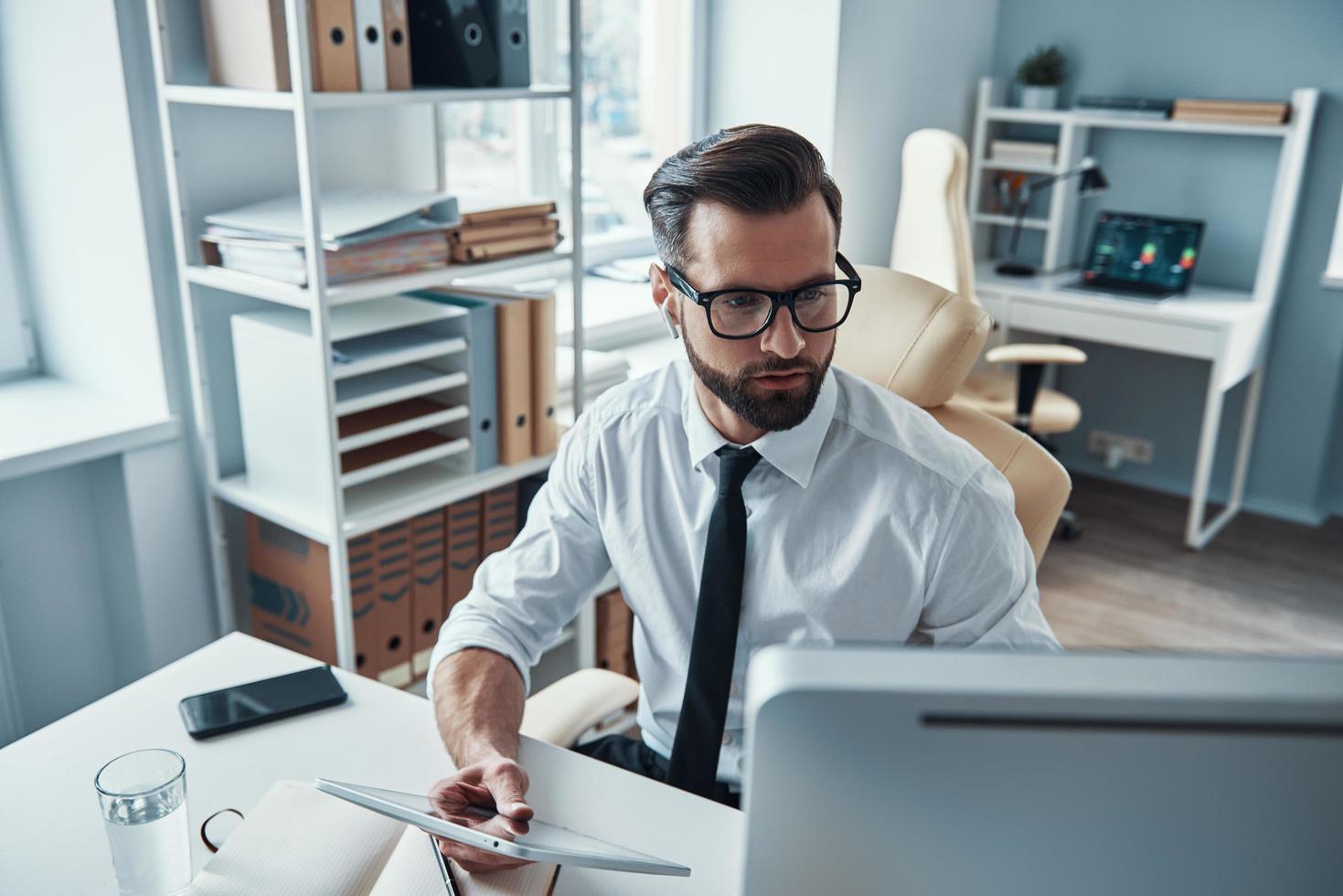 joven concentrado con camisa y corbata trabajando con tecnologías inalámbricas mientras está sentado en la oficina foto