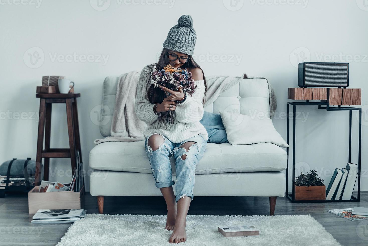 Moment of enjoyment.  Attractive young woman in casual wear and knit hat smelling bouquet and smiling while sitting on the sofa at home photo