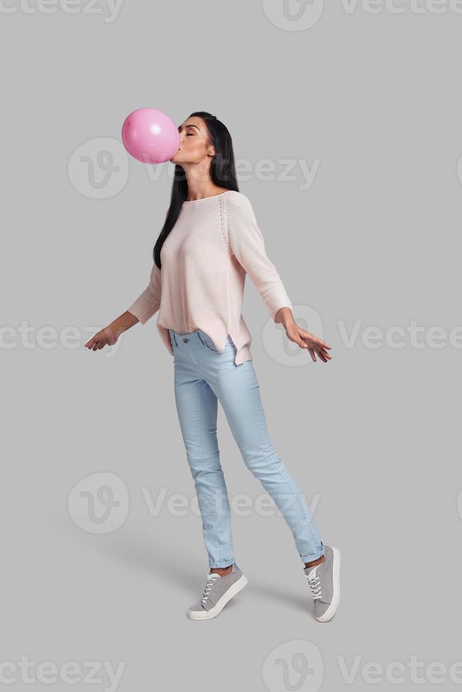 One more step and she will fly. Full length studio shot of attractive young woman in casual wear blowing up a pink balloon while standing against grey background photo