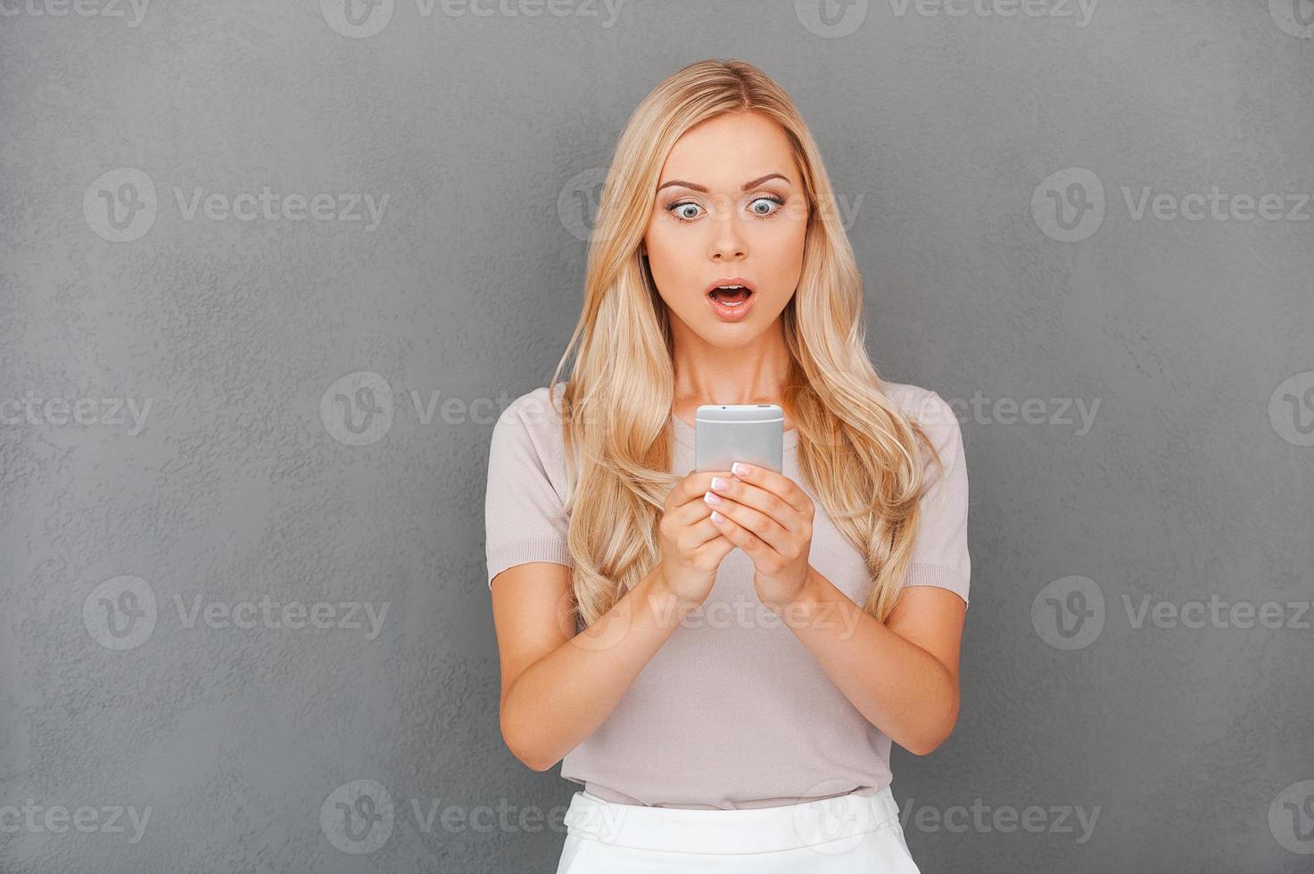 Shocking message. Surprised young blond hair woman holding mobile phone and staring at it while standing against grey background photo
