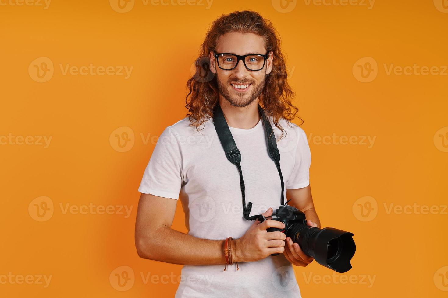 Confident young man in casual clothing looking at camera and holding camera while standing against yellow background photo