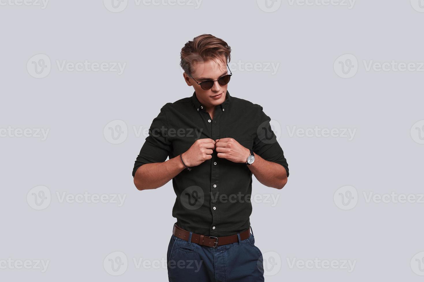 Perfect style. Good looking young man in eyewear buttoning his shirt while standing against grey background photo