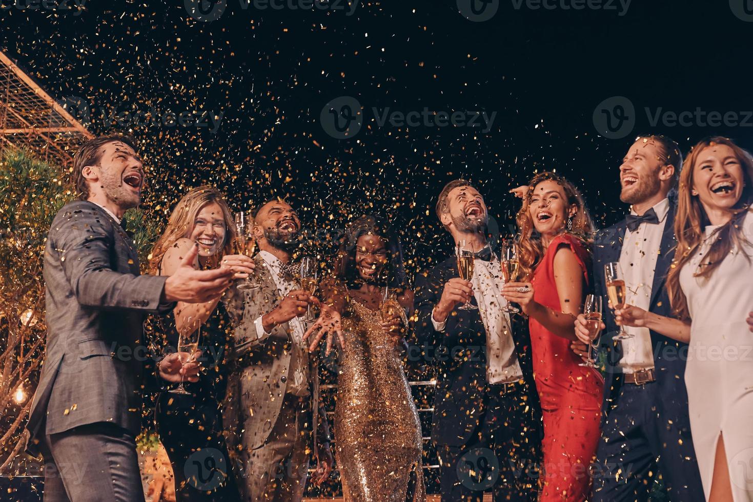 Group of happy people in formalwear having fun together with confetti flying all around photo