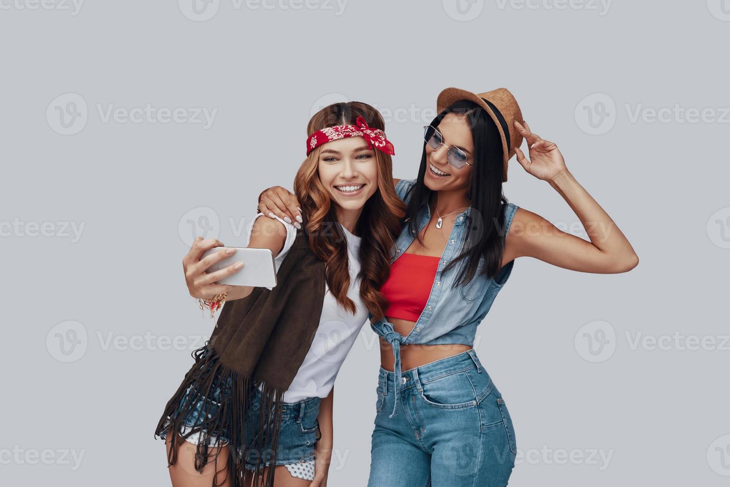 Two attractive stylish young women taking selfie and smiling while standing against grey background photo