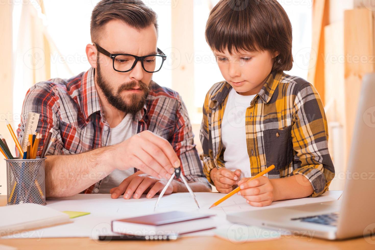 Turning their dreams to reality. Concentrated young man using compass for drawing while sitting at his workplace with his son photo