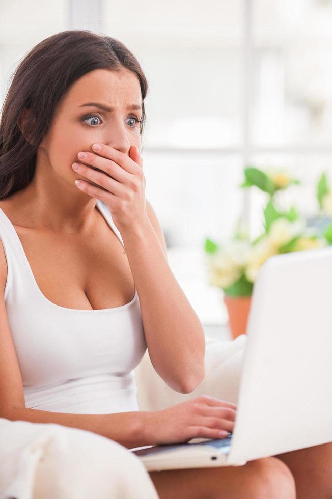 noticias impactantes. mujer joven sorprendida con la mano en la boca y mirando la computadora portátil mientras se sienta en el sofá foto