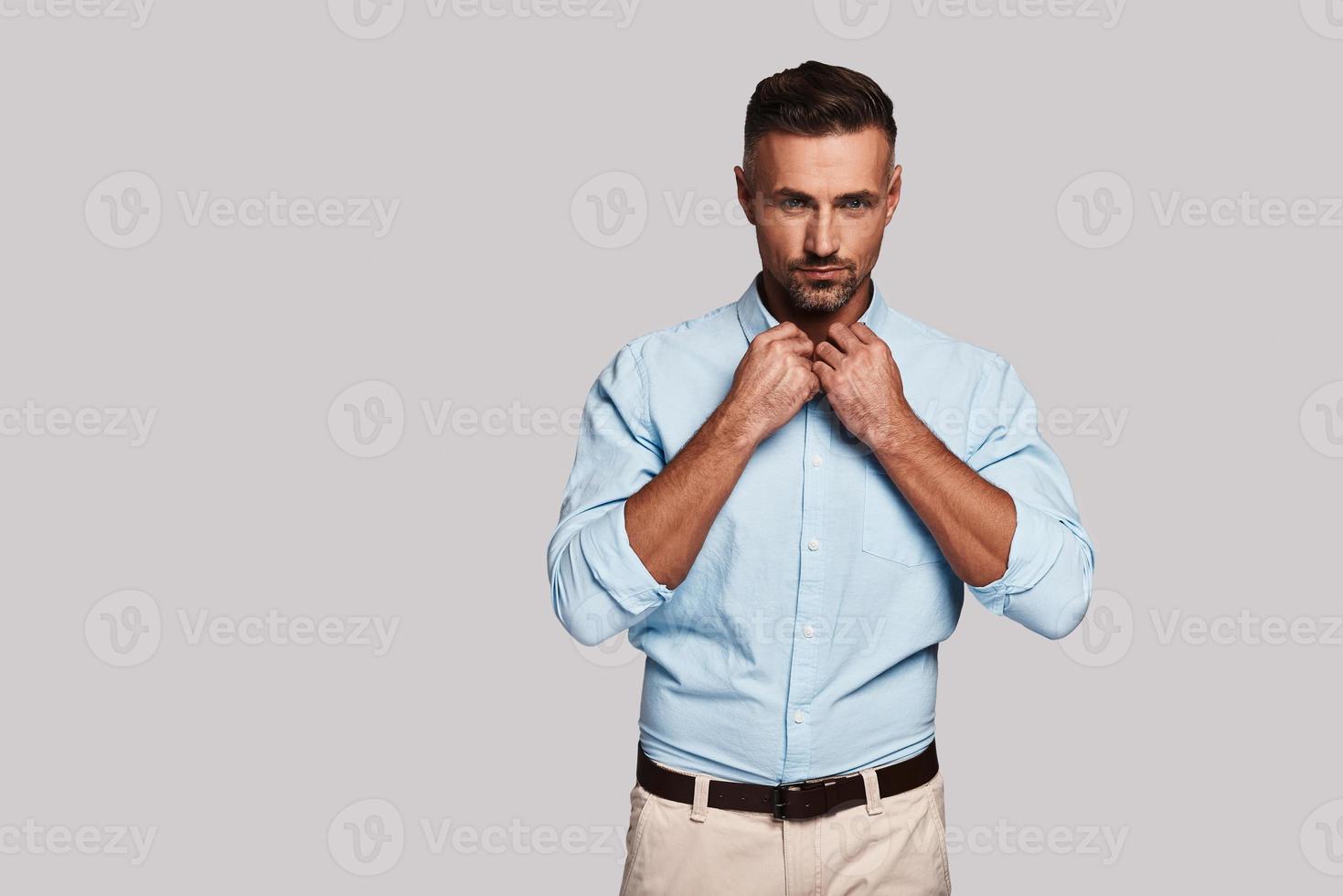 Great sense of style. Handsome young man adjusting collar and looking at camera while standing against grey background photo