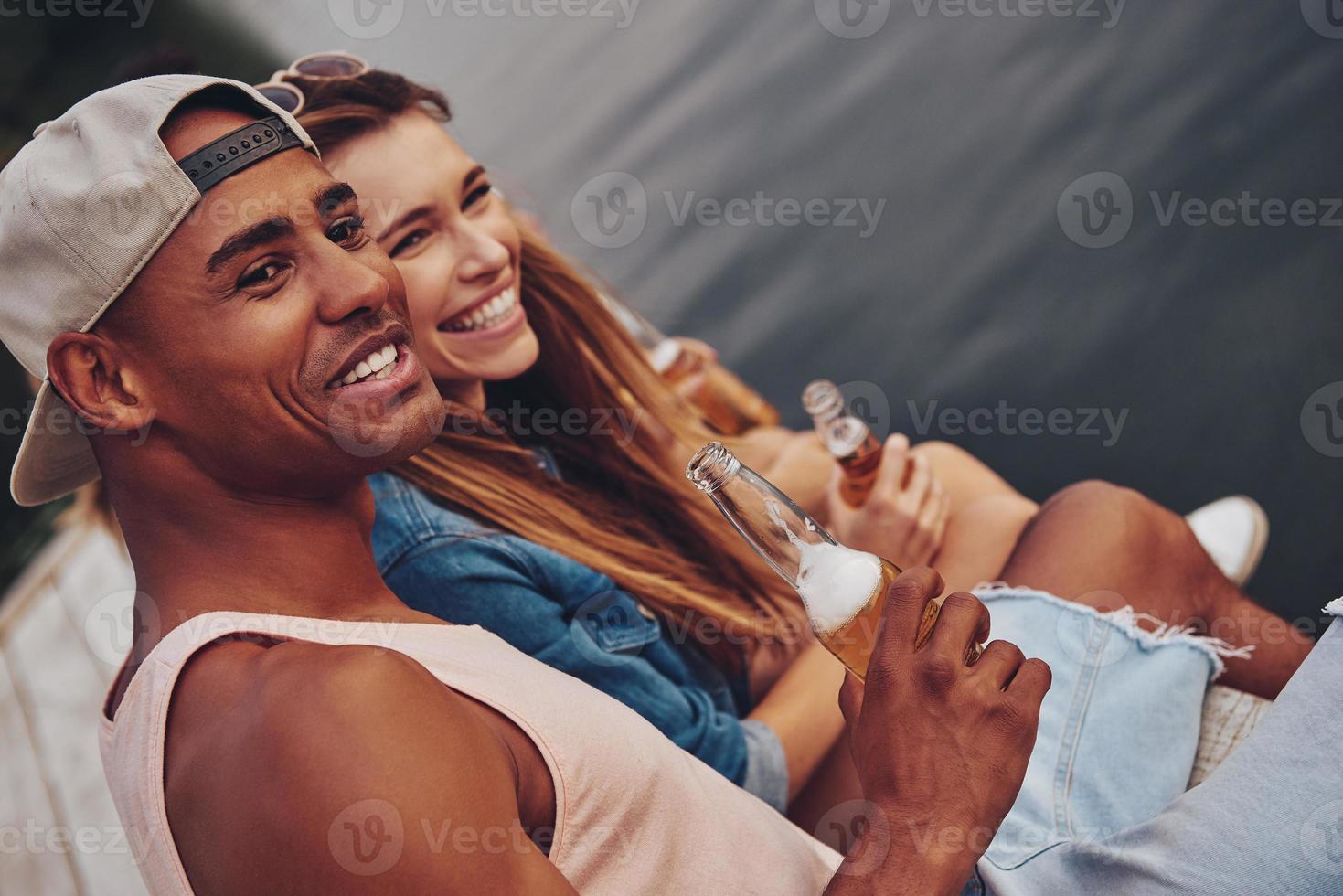 Happy to be around. Group of happy young people in casual wear smiling and drinking beer while sitting on the pier photo