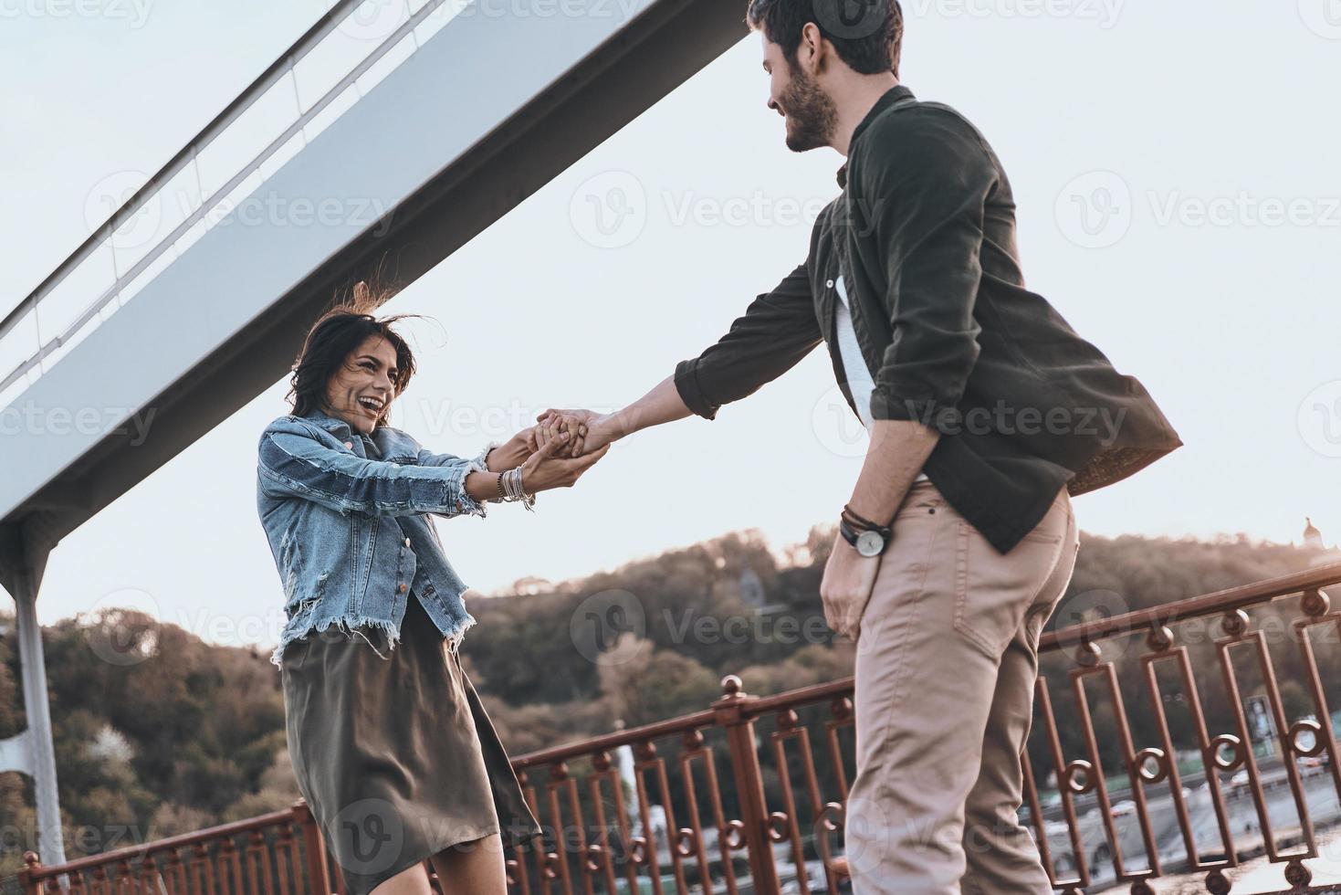 They belong together. Beautiful young couple holding hands and spinning while standing on the bridge outdoors photo