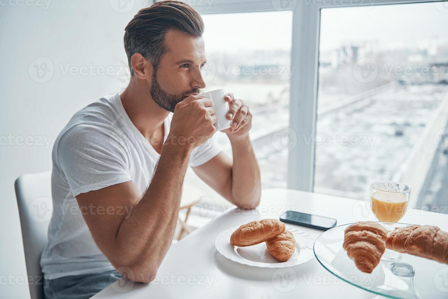 Handsome young man enjoying hot coffee while sitting at the kitchen photo