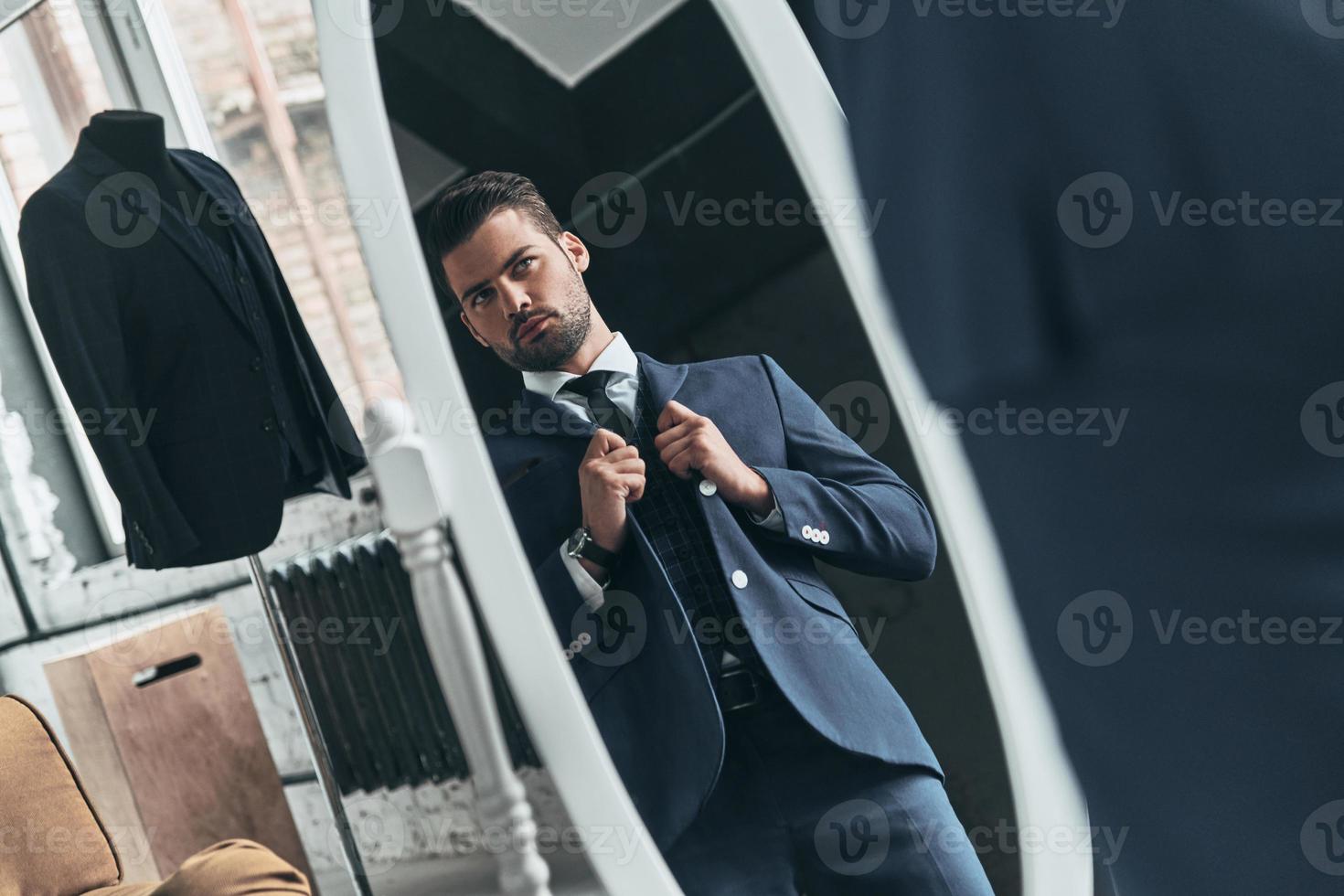 Confident in his style. Reflection of handsome young man in full suit adjusting his jacket while standing in front of the mirror indoors photo