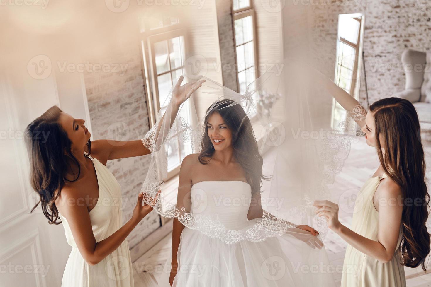 Enjoying wedding preparations. Top view of bridesmaids helping bride to put on a veil and smiling while standing in the fitting room photo
