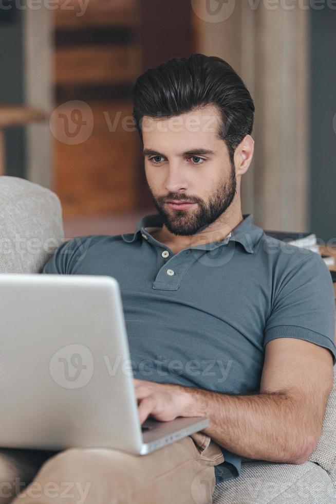 Modern blogger. Handsome young man using his laptop while sitting on the couch at home photo