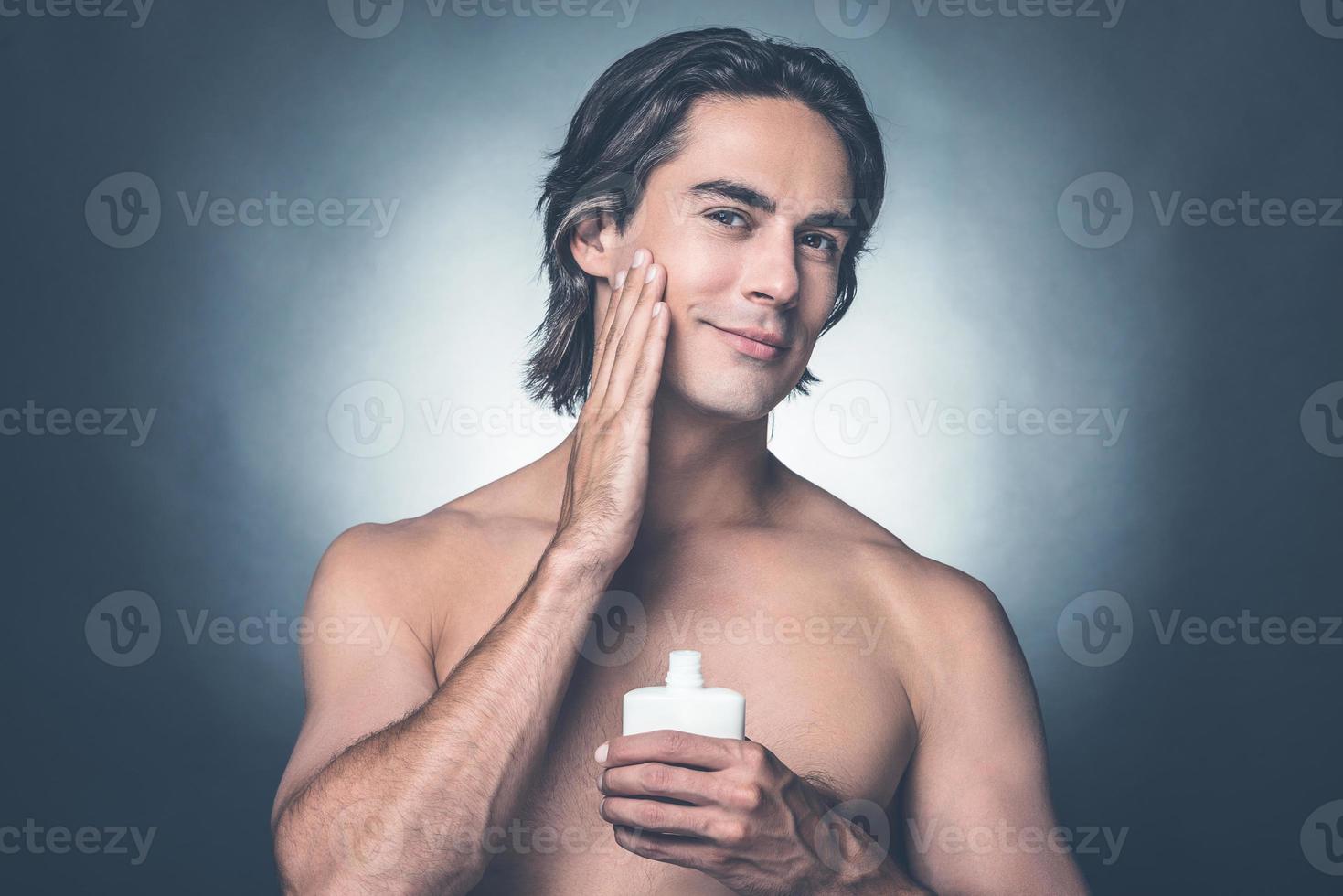Feeling fresh and clean. Portrait of handsome young shirtless man looking at camera and applying aftershave lotion on face while standing against grey background photo