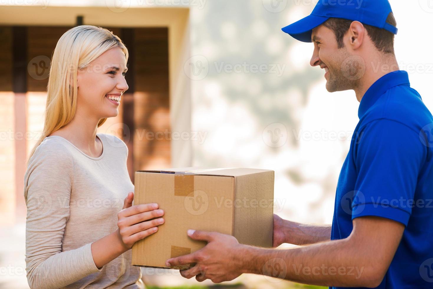 siempre llega a tiempo. vista lateral del joven repartidor feliz dando una caja de cartón a la mujer joven mientras está de pie frente a la casa foto