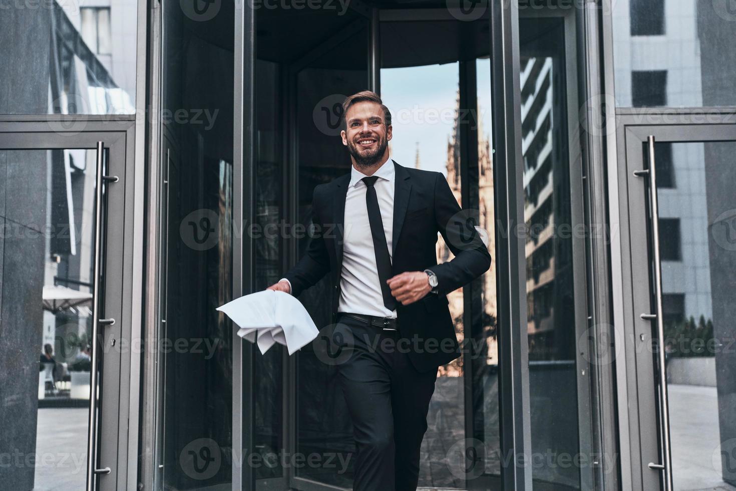 apuesto joven hombre de negocios con traje completo sonriendo y sosteniendo un documento mientras camina al aire libre foto