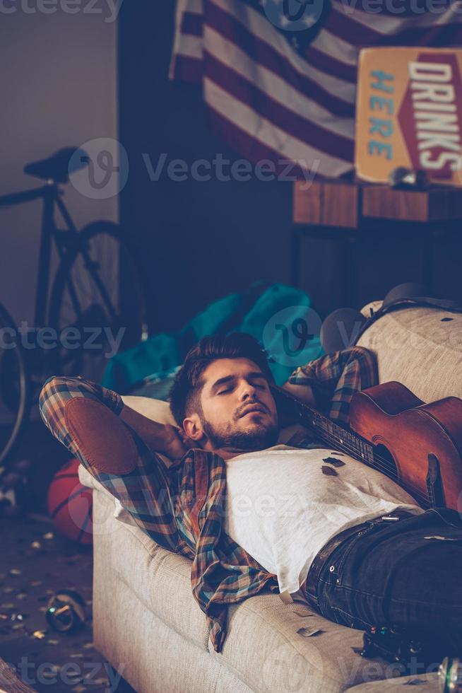 Sleeping off that hangover. Young handsome man lying down on sofa and napping in messy room after party photo