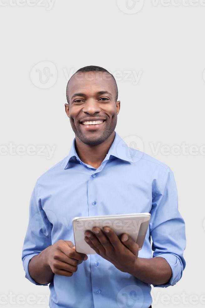Working on digital tablet. Front view of handsome African man working on digital tablet and smiling at camera while standing isolated on grey photo
