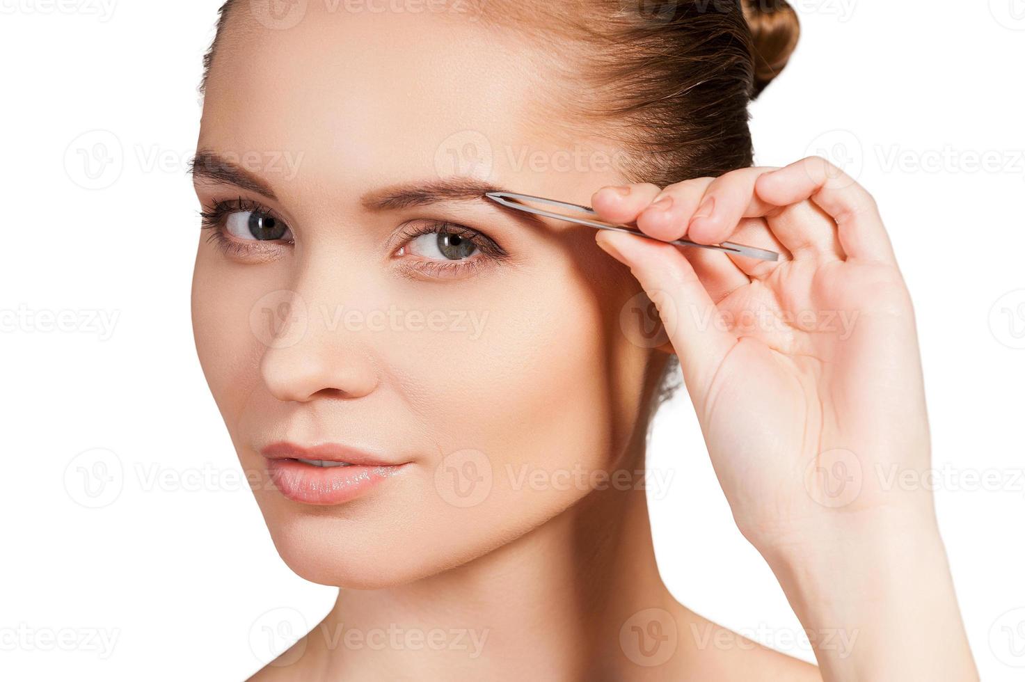 Tweezing her eyebrows. Beautiful young shirtless woman tweezing her eyebrows and looking at camera while standing isolated on white background photo