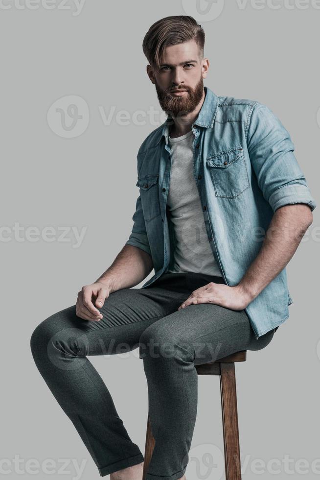 mirada mágica. joven hipster elegantemente vestido mirando a la cámara mientras se sienta en una silla con fondo gris foto