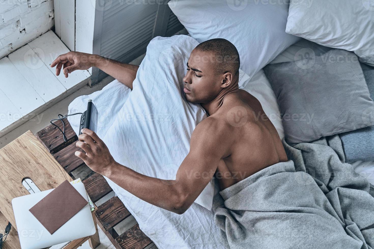 Too early. Top view of young African man turning off the alarm clock while lying in the bed at home photo