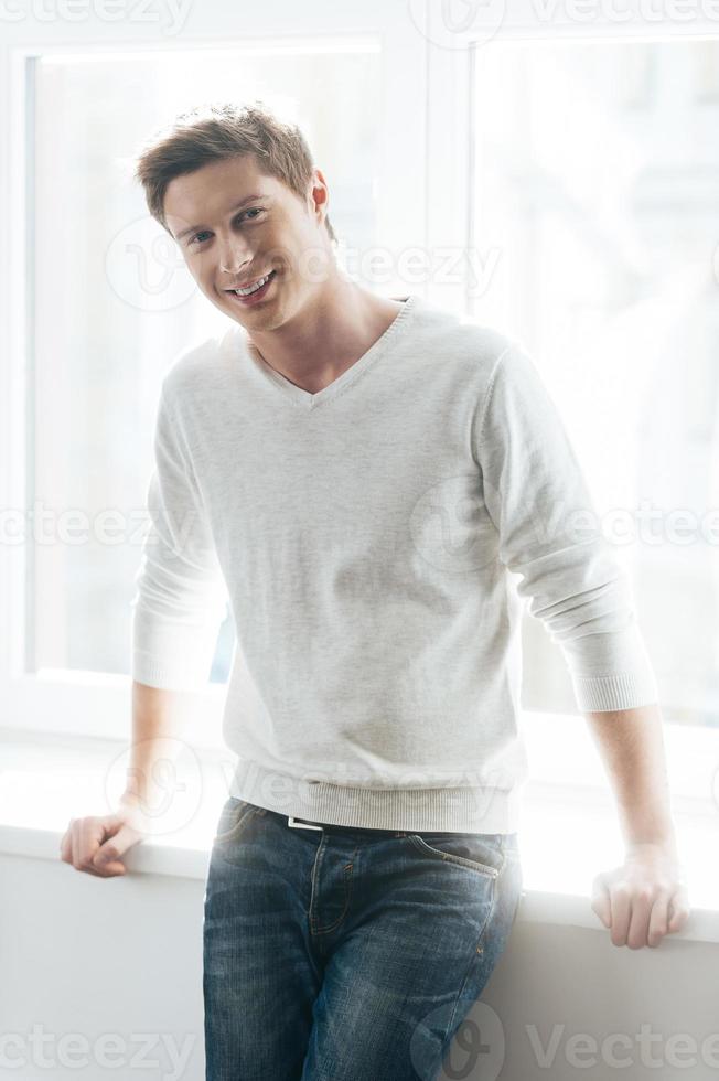 casualmente guapo. apuesto joven apoyado en el alféizar de la ventana y sonriendo foto