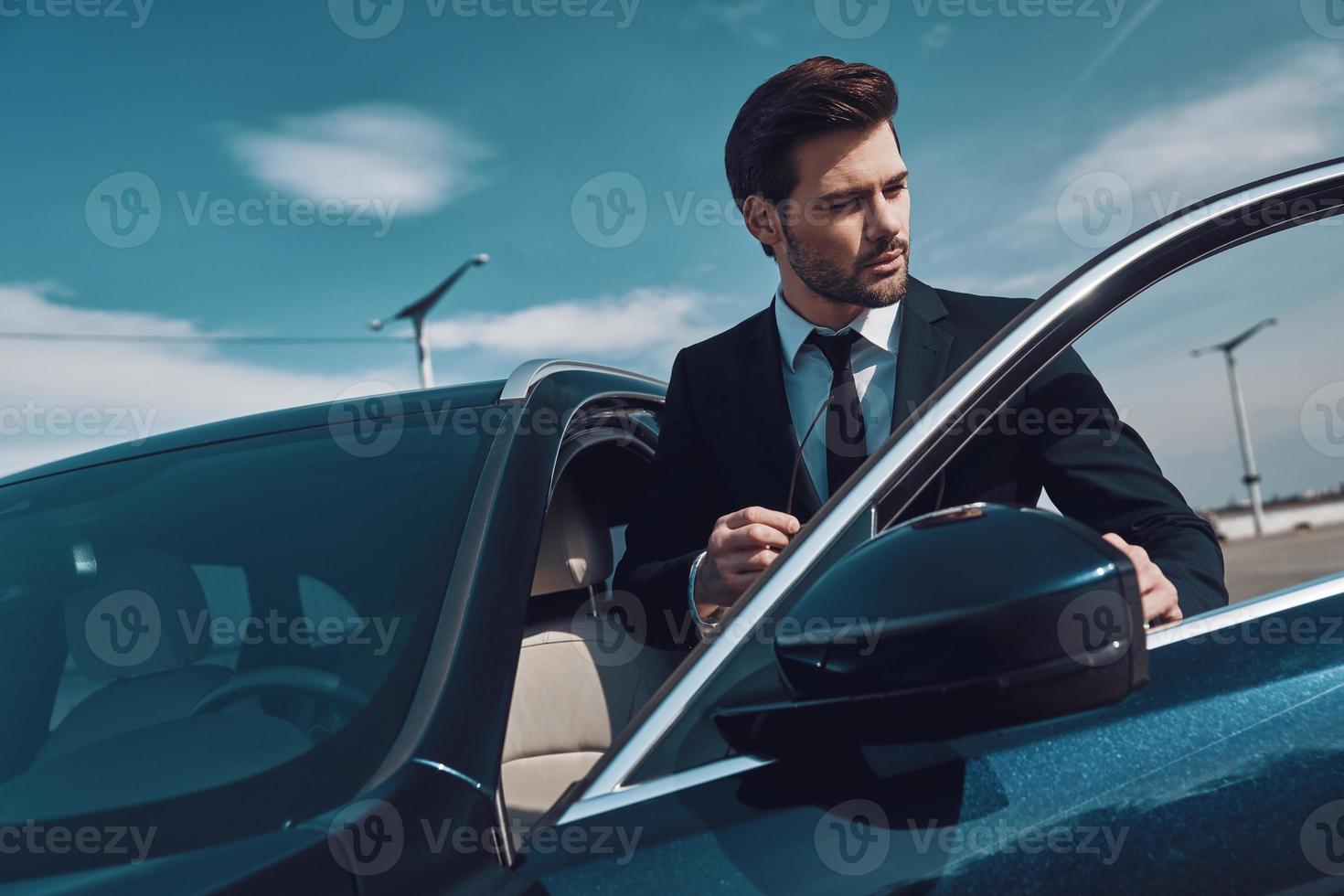 viajes de negocios apuesto joven en ropa de negocios entrando en su coche mientras está de pie al aire libre foto