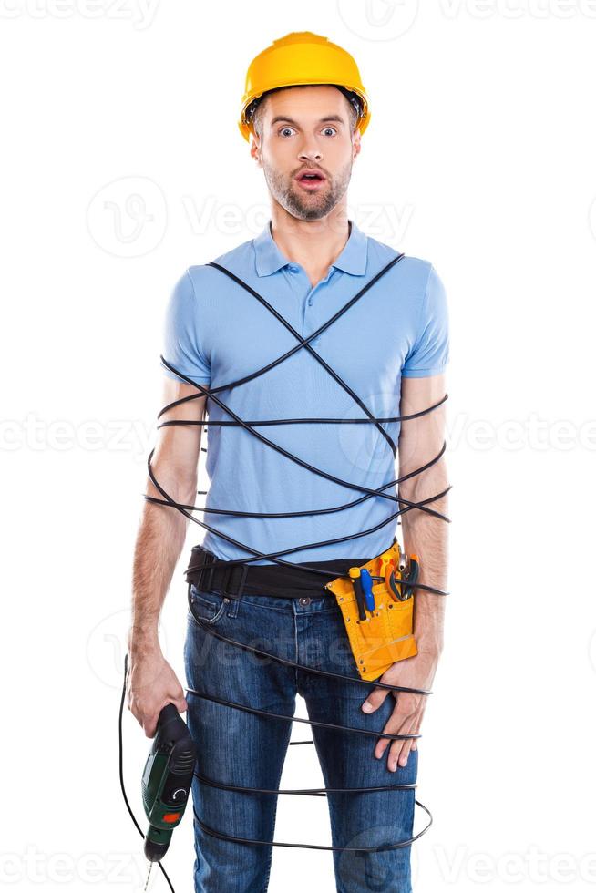 Frustrated carpenter. Frustrated young male carpenter trapped in wire holding drill and keeping mouth open while standing against white background photo