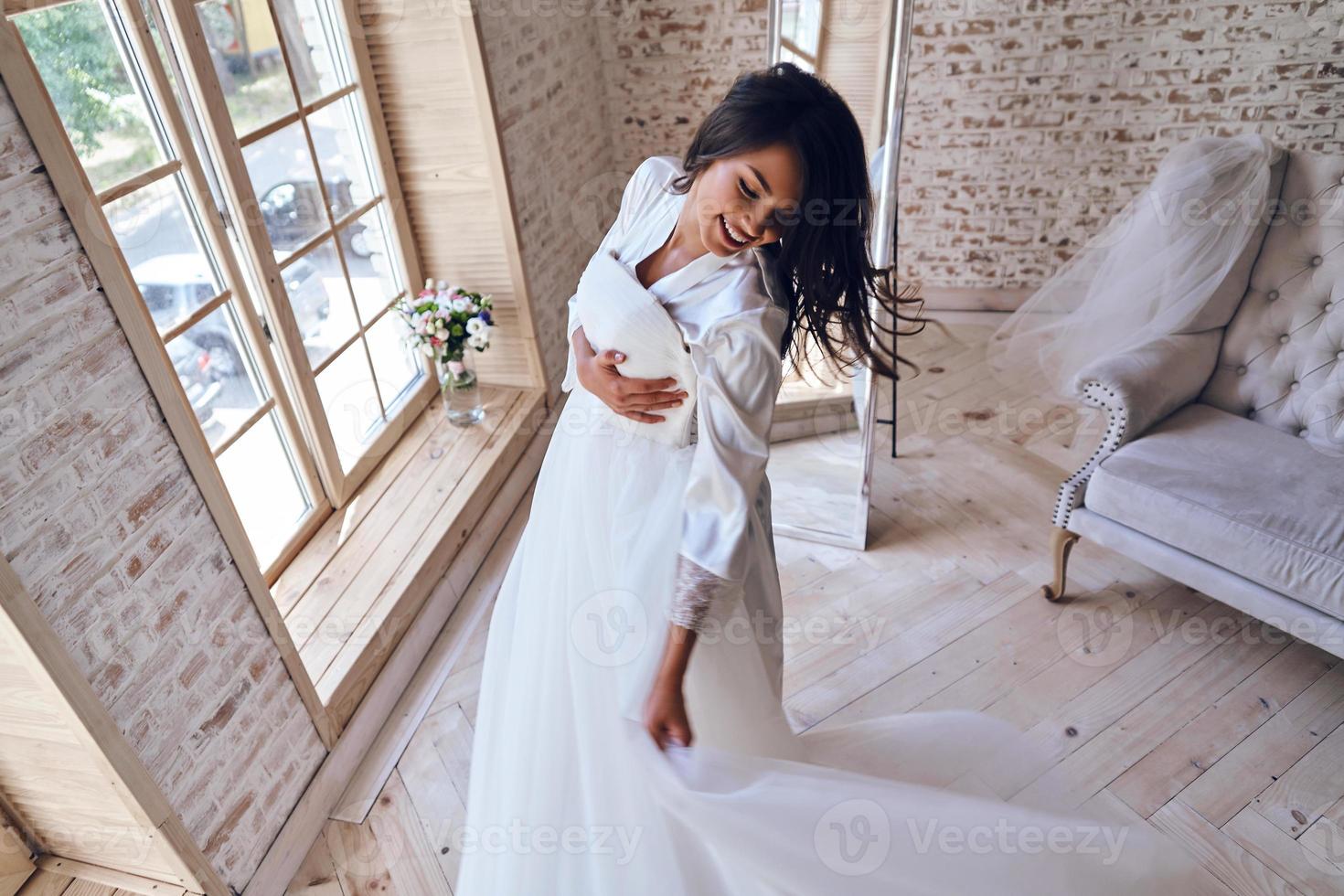 disfrutando de su día especial. vista superior de una atractiva joven con albornoz de seda probándose su vestido de novia y sonriendo mientras está de pie cerca de la ventana foto