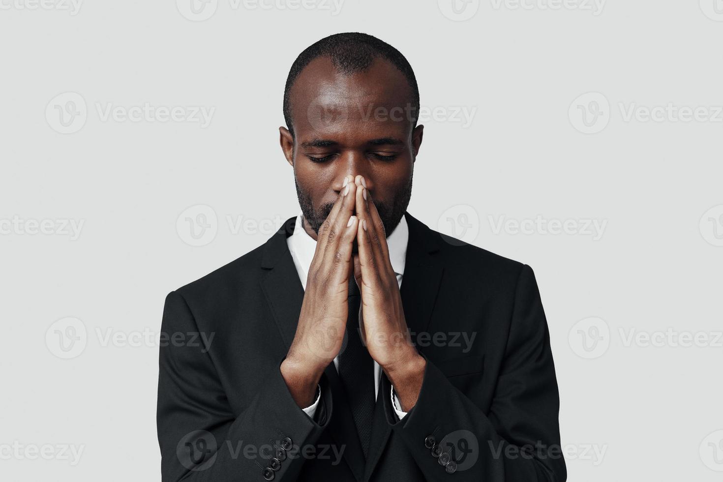 Young African man in formalwear keeping hands clasped while standing against grey background photo