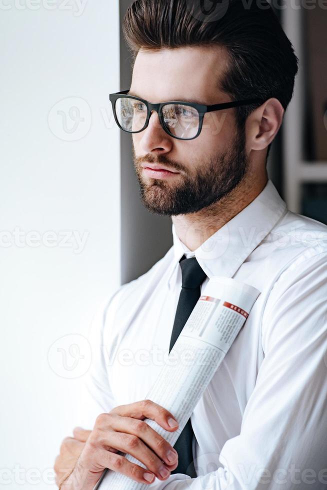 analizando las noticias de los medios. un joven hombre de negocios pensativo con anteojos sosteniendo un periódico enrollado y mirando hacia otro lado mientras estaba en el cargo foto