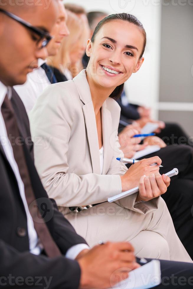 Businesswoman at the conference. Side view of business people sitting in a row and writing something in their note pads while confident young woman looking at camera and smiling photo