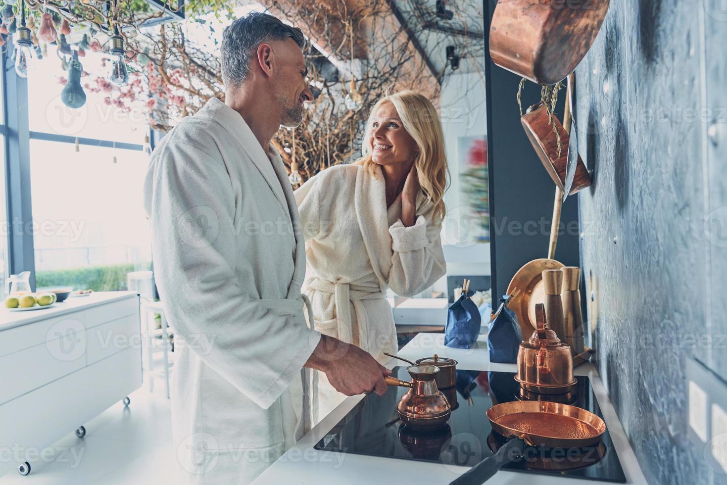 hermosa pareja madura en albornoces preparando café juntos mientras pasan tiempo en la cocina doméstica foto