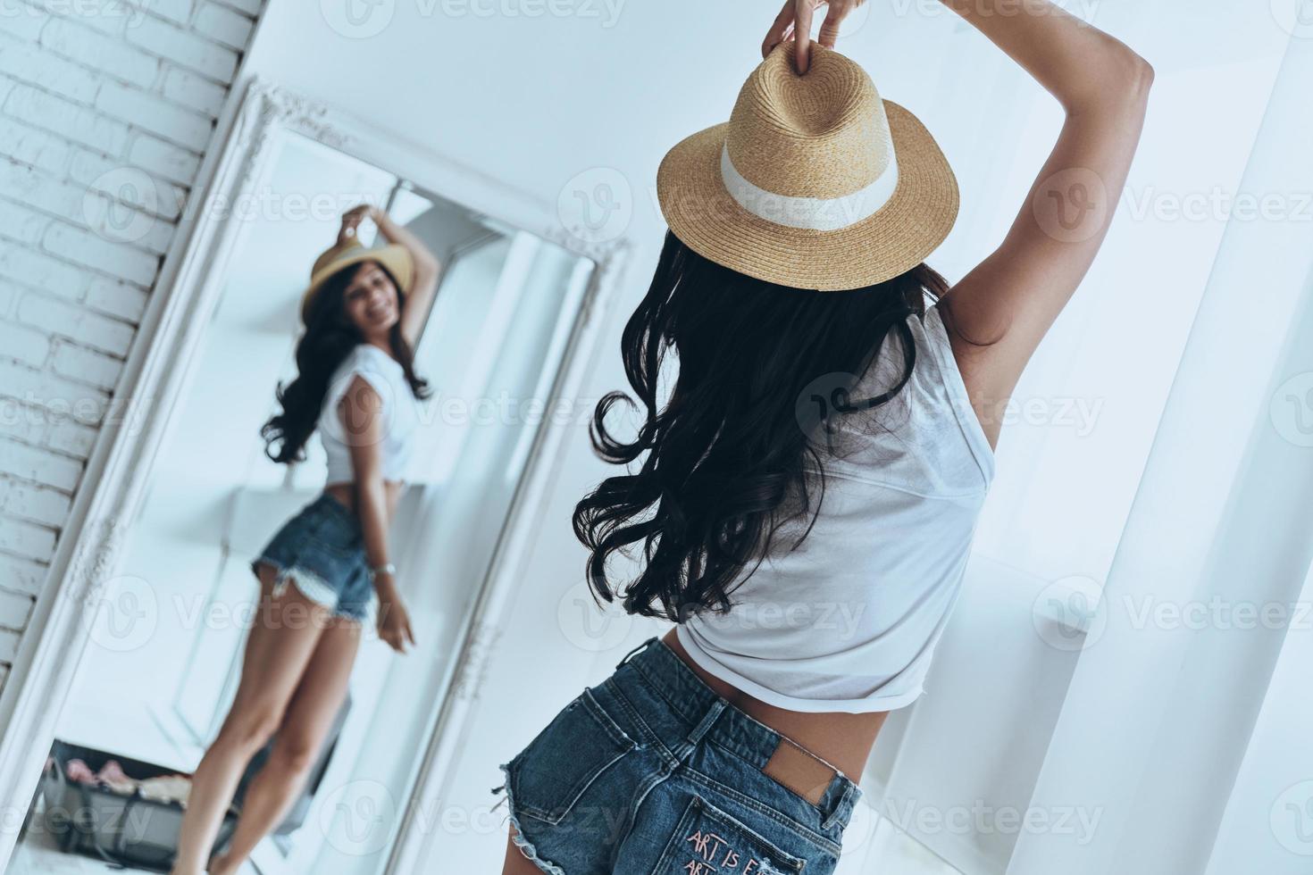 Ready to vacation. Rear view of attractive young woman trying on her sun hat and smiling while looking in the mirror at home photo