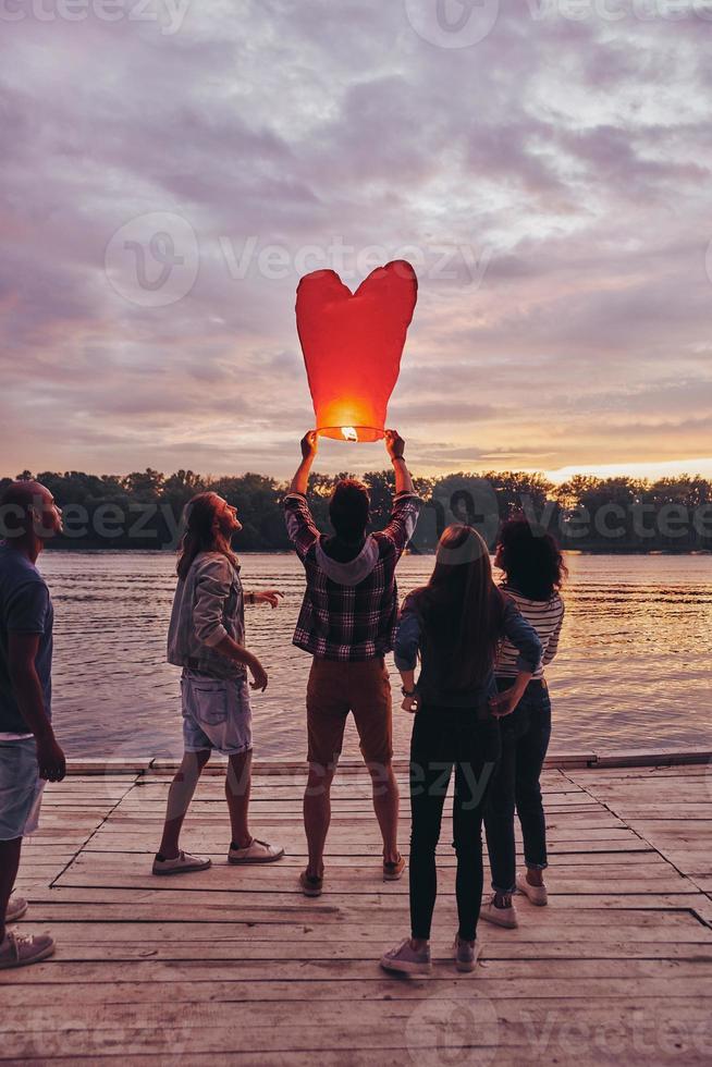 Carefree and happy. Full length of young people in casual wear preparing sky lantern while standing on the pier photo