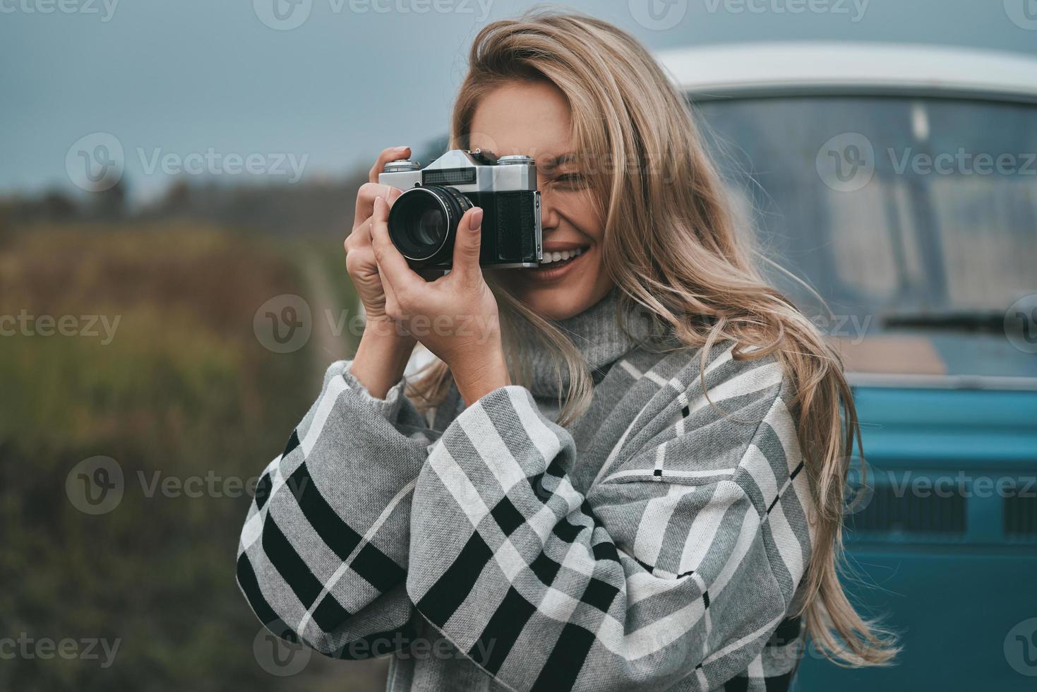 Cool Shot atractiva mujer joven fotografiando y sonriendo mientras está de pie al aire libre con la mini furgoneta de estilo retro azul en el fondo foto