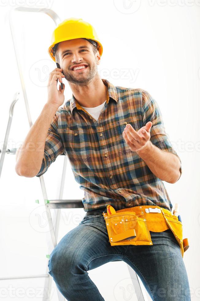 descanso para una charla encantadora. alegre joven reparador hablando por teléfono móvil mientras está sentado en la escalera foto