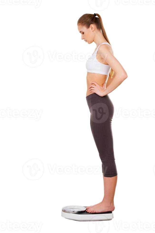 Woman on weight scale. Side view of beautiful young woman in sports clothing standing on weight scale and looking down photo