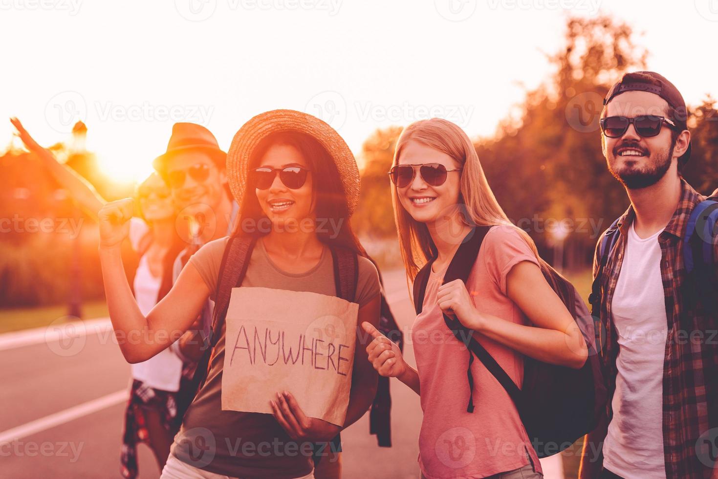 Destination is anywhere. Beautiful young people with backpacks hitchhiking on the road and looking happy photo