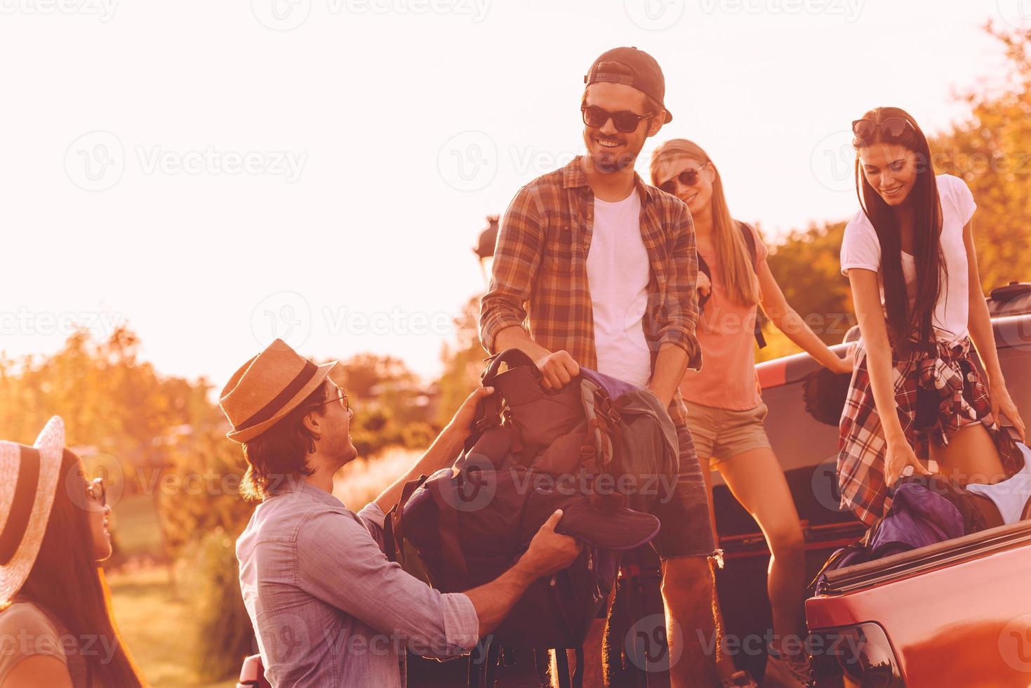 Loading backpacks to truck. Beautiful young people loading their backpacks to pick-up truck and looking happy with sunset in the backgrounds photo
