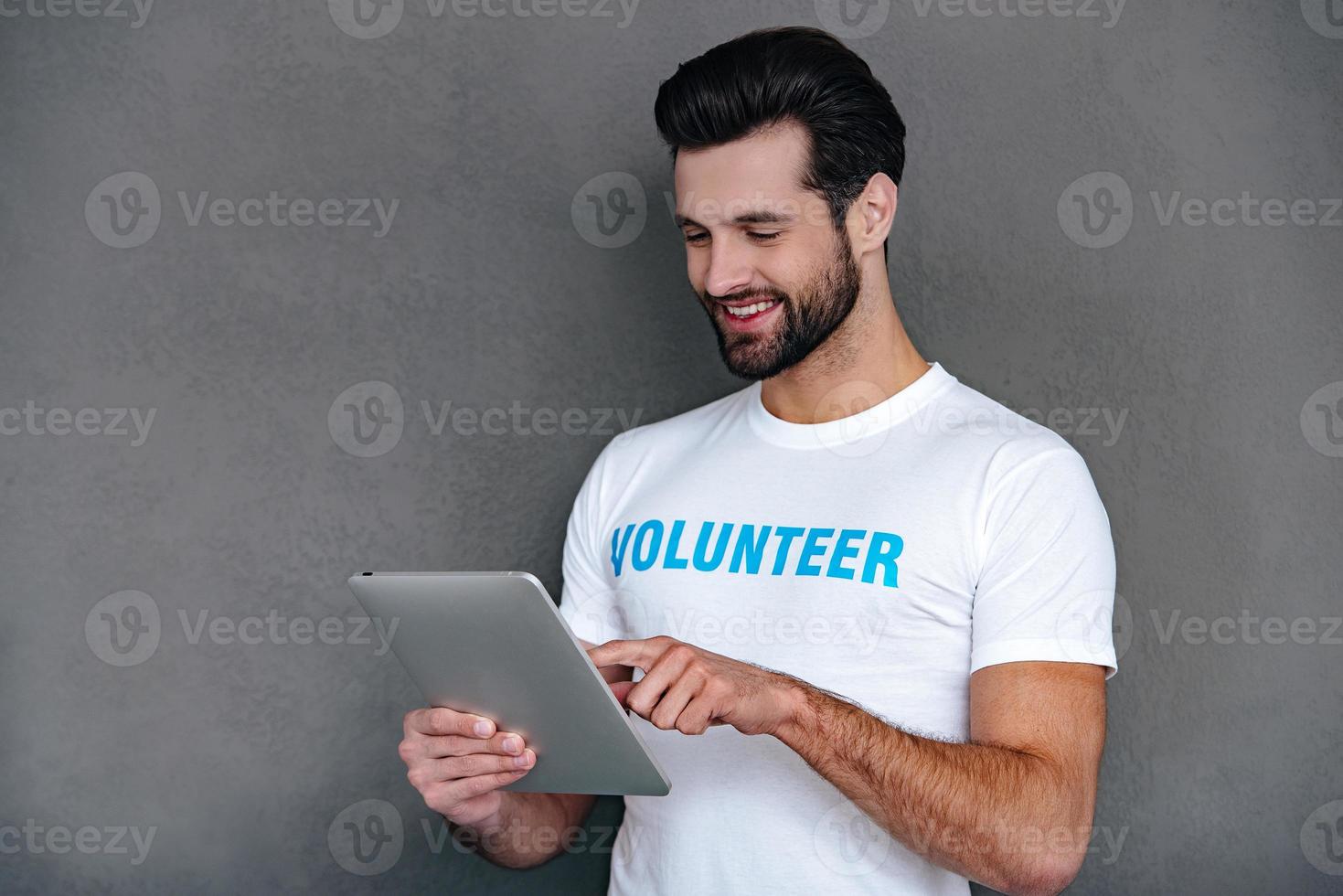 Donation by one click. Confident young man in volunteer t-shirt using his digital tablet with smile while standing against grey background photo